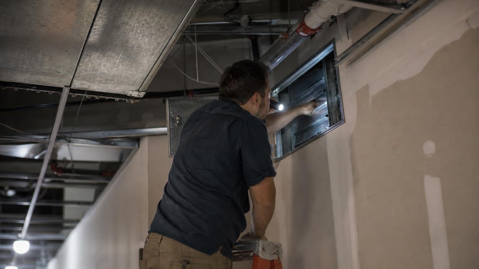 Technician inspecting a fire damper through a duct access door above a corridor ceiling